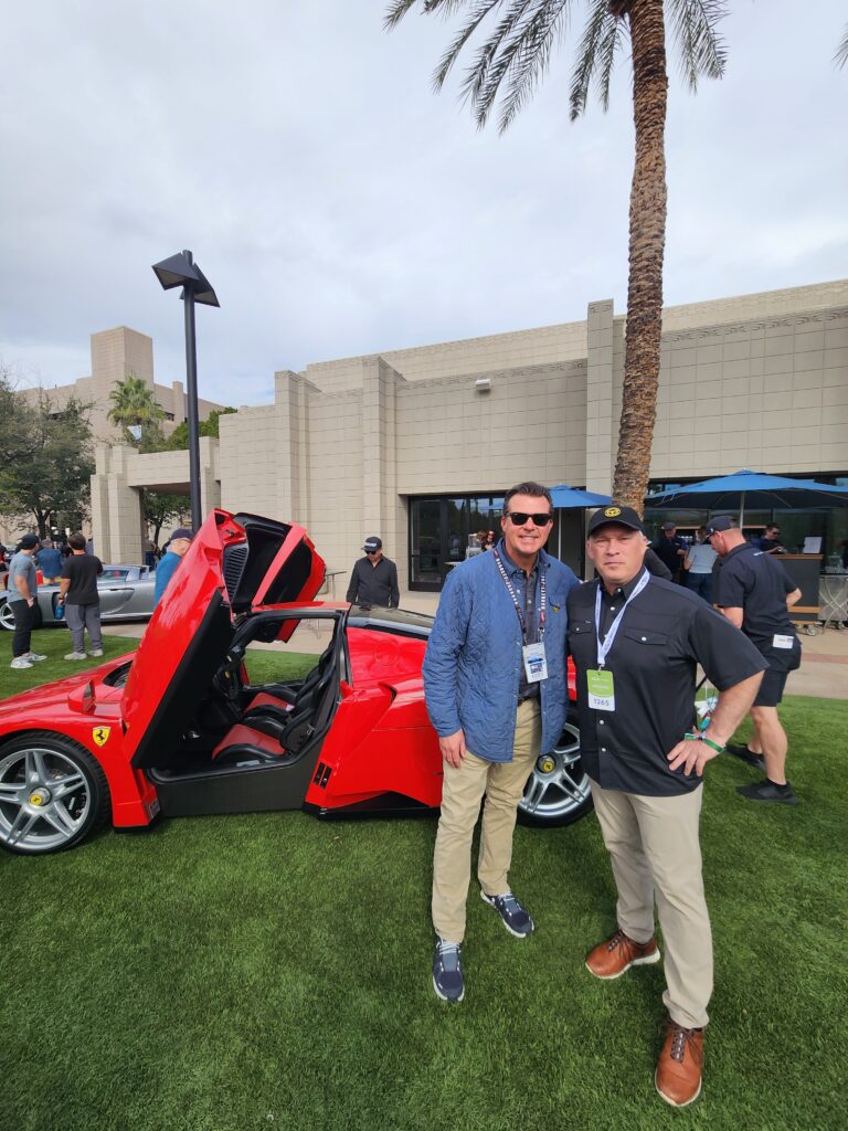 Two men posing beside a red sports car with open doors at an outdoor event.